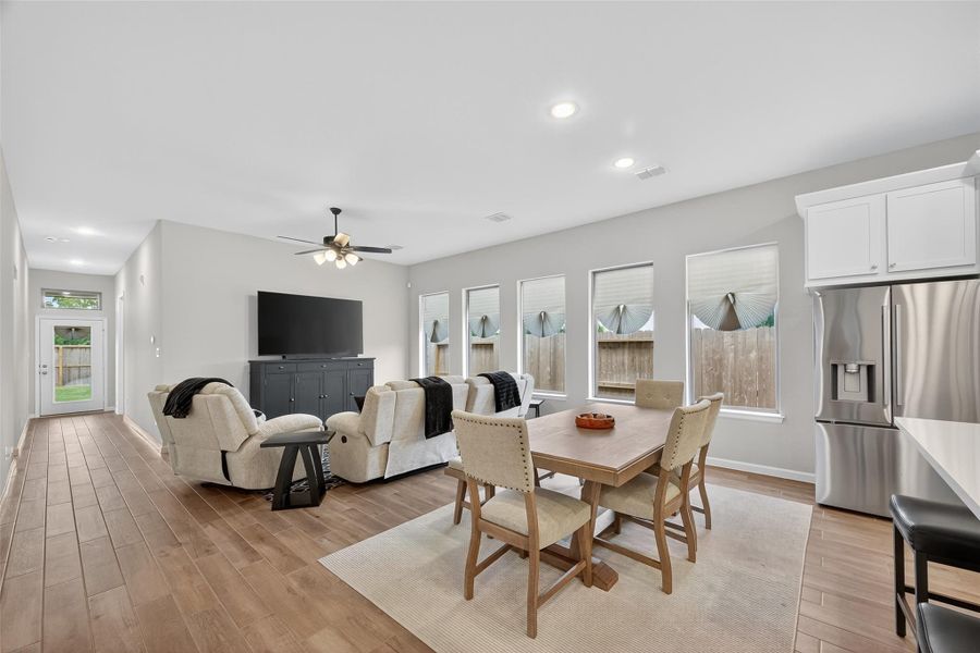 This spacious open-concept living area is dressed with a cozy seating arrangement and a dining table for four. The room is well-lit with large windows, modern ceiling fans, and recessed lighting. The kitchen area includes sleek stainless steel appliances and a breakfast bar, with wood look flooring throughout. This spacious open-concept living area is dressed with a cozy seating arrangement and a dining table for four. The room is well-lit with large windows, modern ceiling fans, and recessed lighting. The kitchen area includes sleek stainless steel appliances and a breakfast bar, with wood look flooring throughout.