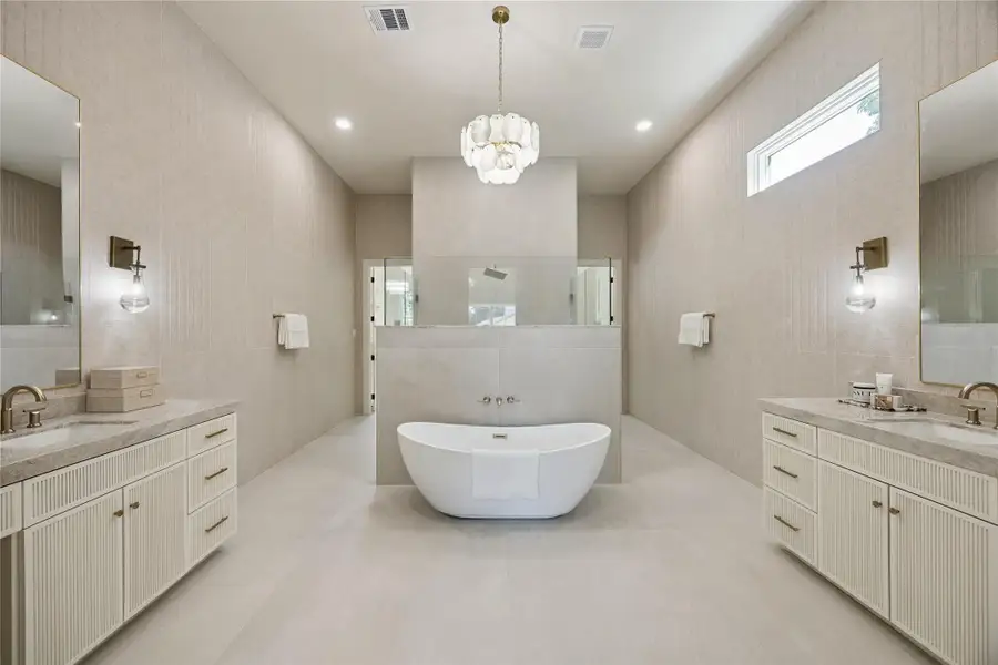 PRIMARY BATH: Spa-inspired primary bath offers separate vanities with Taj Mahal Quartzite countertops, Capri Stone from Porcelanosa, a freestanding soaking tub, and an oversized glass-enclosed shower with body sprayers.