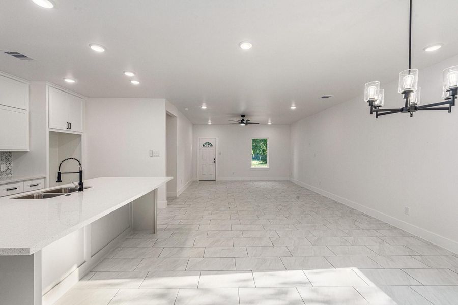 This spacious open-concept living area features modern white cabinetry, a sleek kitchen island with a built-in sink, and elegant lighting fixtures. The room is bright with ample natural light and has a clean, contemporary tile floor.