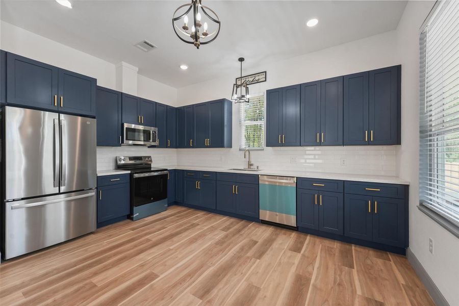 This kitchen features sleek blue cabinets with gold hardware, stainless steel appliances, and a stylish chandelier. The space is bright, with a large window and wood-look flooring, creating a modern and inviting atmosphere.
