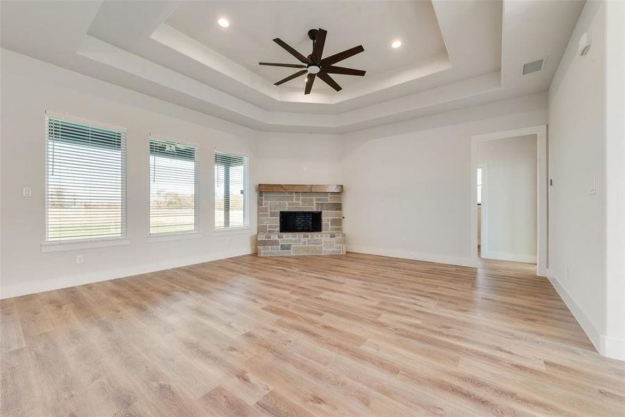 Spacious room featuring wood-finish flooring, a stone-faced fireplace with a wood mantle, and a multi-tiered tray ceiling with a dark-bladed ceiling fan