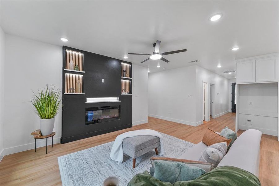 Living area featuring a contemporary dark accent wall with an electric fireplace, integrated shelving with accent lighting, and wood-finish flooring Living area featuring a contemporary dark accent wall with an electric fireplace, integrated shelving with accent lighting, and wood-finish flooring