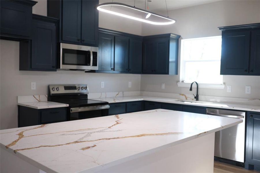 Kitchen featuring appliances with stainless steel finishes, light stone countertops, a kitchen island, and decorative light fixtures Kitchen featuring appliances with stainless steel finishes, light stone countertops, a kitchen island, and decorative light fixtures