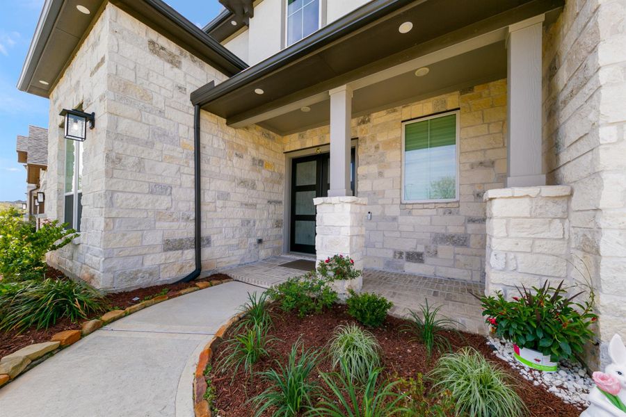 Charming covered front porch with elegant stone façade and inviting entryway, creating a warm first impression. Beautifully landscaped garden beds and a curved walkway add to the home’s curb appeal and welcoming feel.