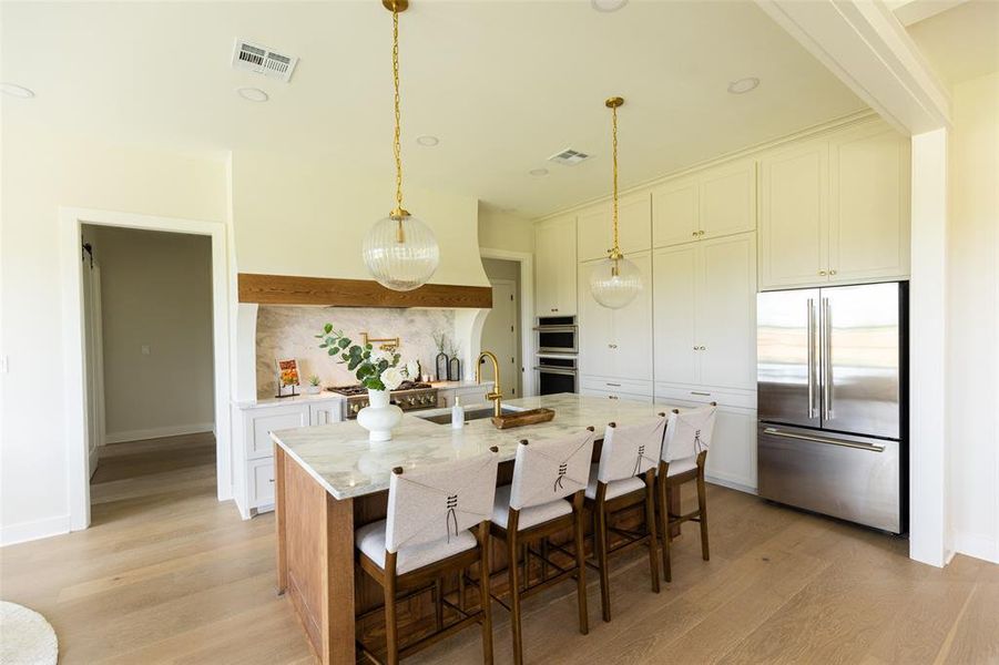 Spacious kitchen featuring a large central island with a stone countertop, light wood-finish flooring, white cabinetry with gold-toned hardware, stainless steel refrigerator, and integrated double ovens