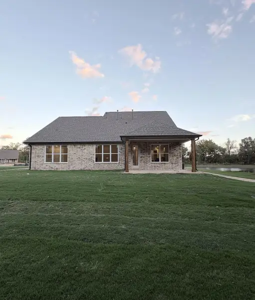 Brick exterior residence featuring a covered rear patio with wooden support beams, multi-pane windows, a pitched shingle roof, and an expansive lawn