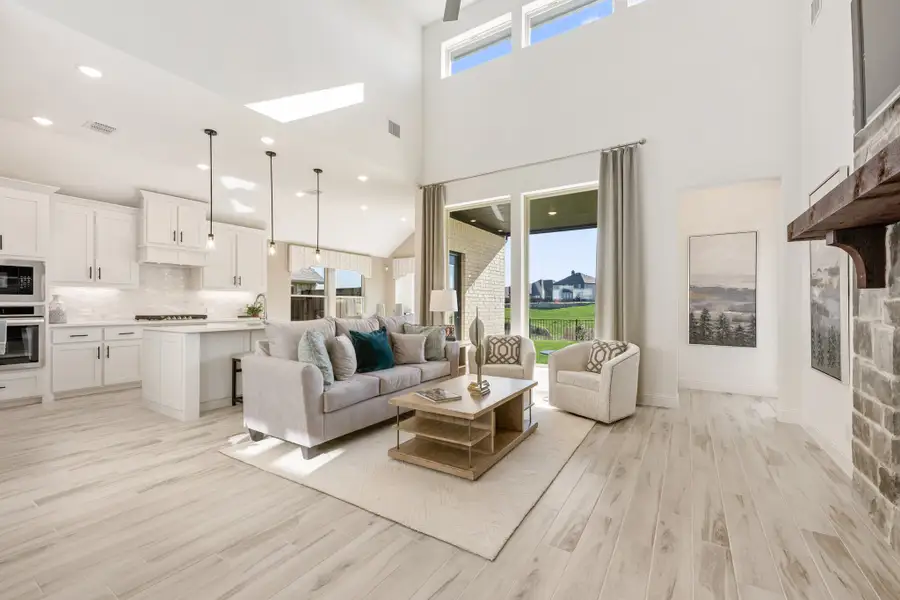 Open-concept living room with two-story ceiling, light wood floors, and kitchen visible in background