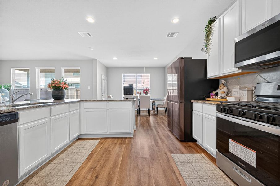 This kitchen features modern white cabinetry, granite countertops, and stainless steel appliances, including a gas stove. The open layout flows into a bright dining area with large windows, and the wood flooring adds warmth to the space.