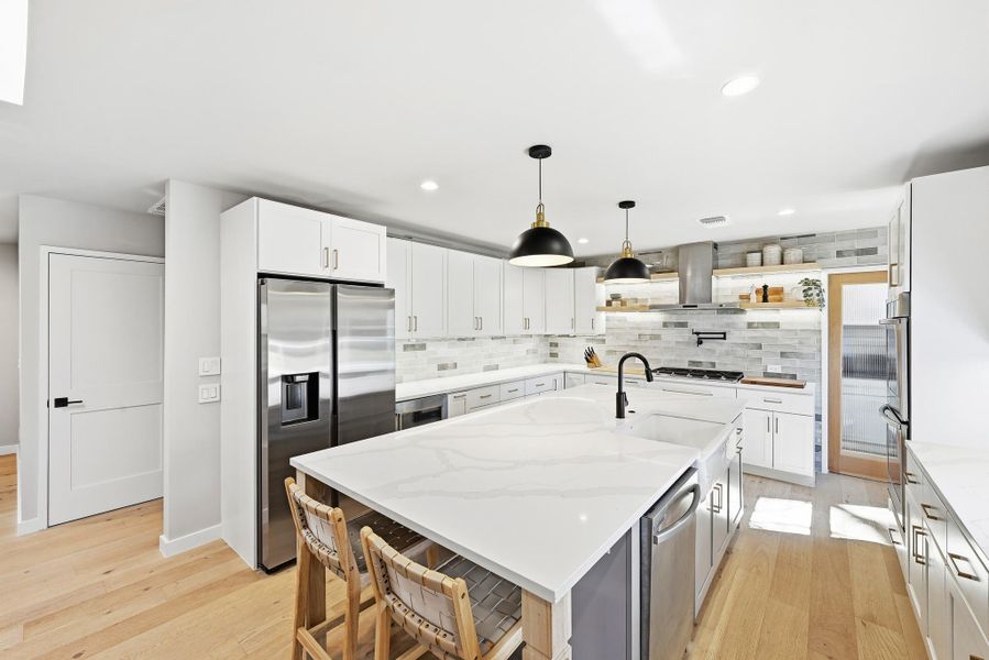 Kitchen with stainless steel appliances, light wood-type flooring, hanging light fixtures, light stone countertops, and a center island with sink