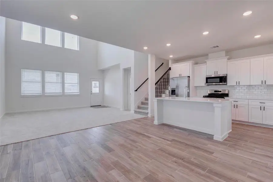 Bright open-concept view connecting the kitchen, breakfast area, family room, and staircase. The white kitchen cabinetry, large island, quartz-style surfaces, and subway tile backsplash create a fresh modern look. The family room features soaring ceilings, generous windows, and soft carpet, offering a comfortable gathering space at the center of the home. Bright open-concept view connecting the kitchen, breakfast area, family room, and staircase. The white kitchen cabinetry, large island, quartz-style surfaces, and subway tile backsplash create a fresh modern look. The family room features soaring ceilings, generous windows, and soft carpet, offering a comfortable gathering space at the center of the home.