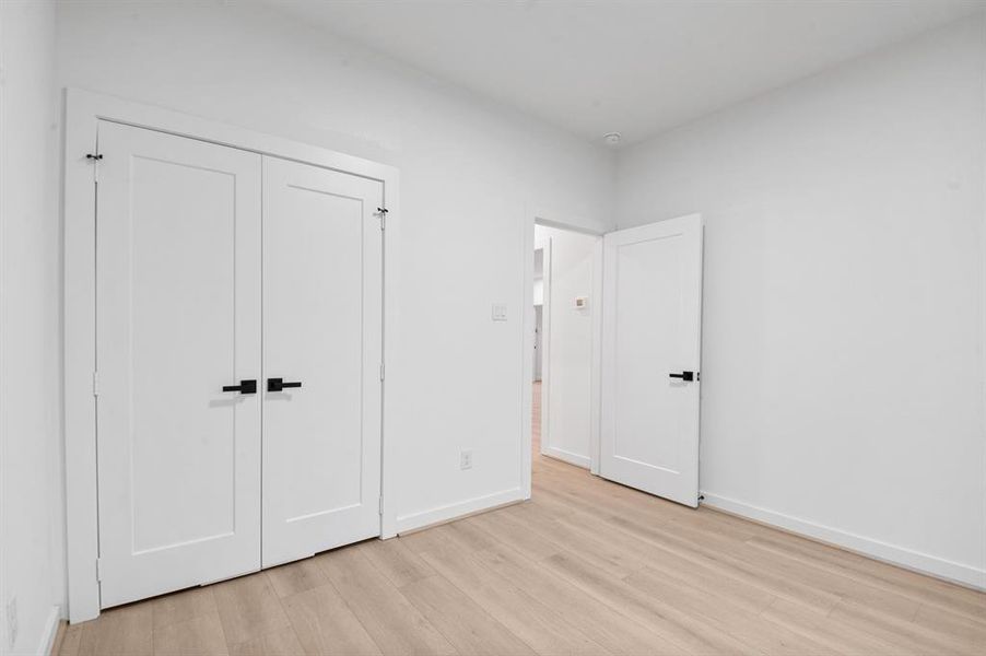 This photo showcases a bright, minimalist room with light wood flooring, white walls, and modern black hardware on the doors. It features a double-door closet and an open entry leading to another area of the home.