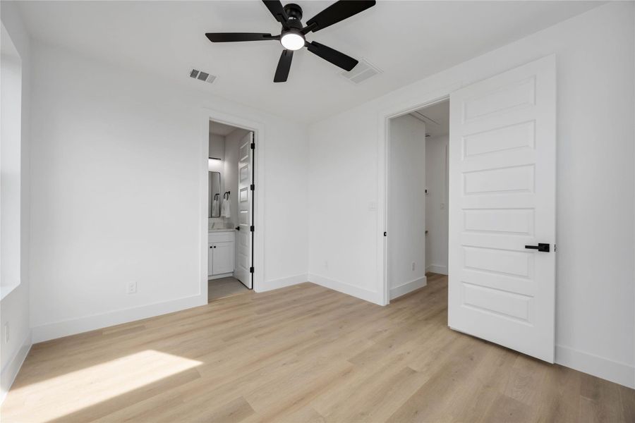 This spacious secondary bedroom features warm wood-look flooring, fresh white walls, and a modern ceiling fan, with direct access to an en-suite bathroom and a convenient adjoining closet for added functionality.
