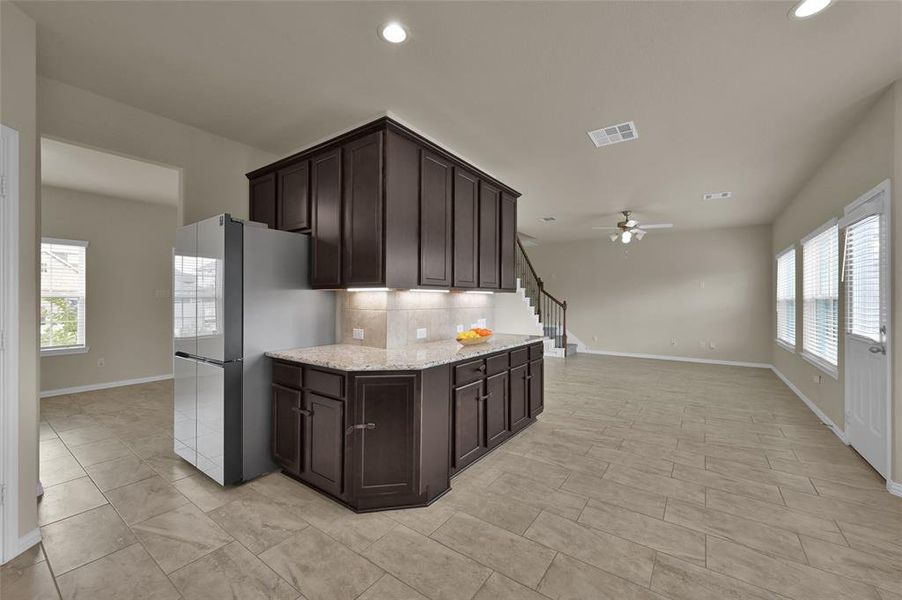 This is a spacious, modern kitchen with dark cabinetry and ample counter space. The open floor plan leads into a well-lit living area with tile flooring and multiple windows.