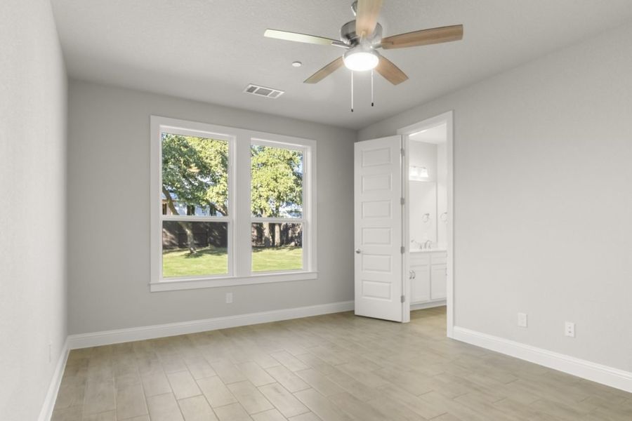 Image of primary bedroom with light grey painted walls, light brown wood-like flooring, two large windows, and a connecting bathroom Image of primary bedroom with light grey painted walls, light brown wood-like flooring, two large windows, and a connecting bathroom