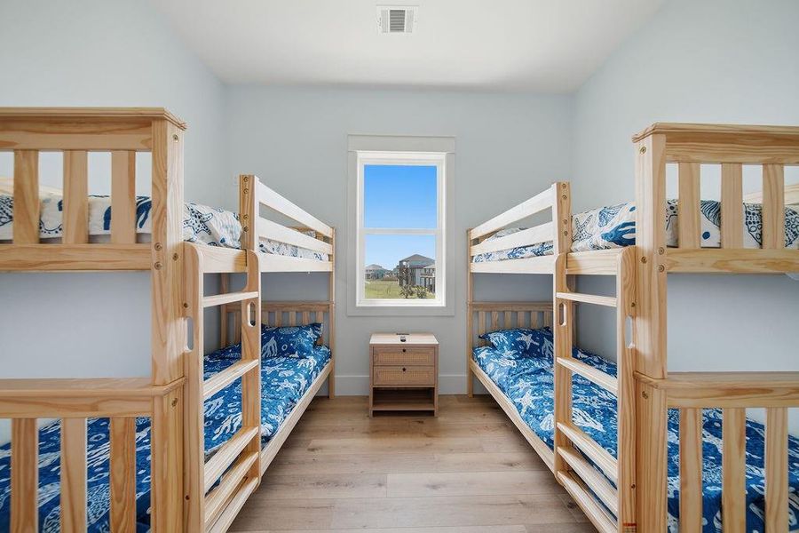 Bright and airy bedroom featuring two sets of wooden bunk beds with blue patterned bedding, a central window offering a view of the outdoors, and a small wooden nightstand. Ideal for accommodating multiple guests or family members.