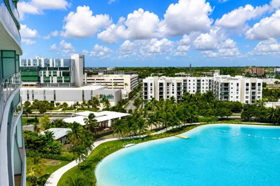 Elevated balcony featuring a glass railing, overlooking a large turquoise lagoon, landscaped grounds, palm trees, and paved walking paths