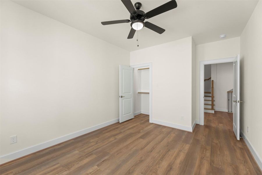 This room features a clean, modern design with light walls and flooring. It includes a ceiling fan, a small closet with an open door, and an adjacent door leading to a hallway with a staircase.