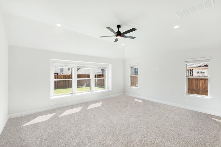 Empty bedroom with vaulted ceiling, ceiling fan, carpet flooring, and large windows overlooking backyard