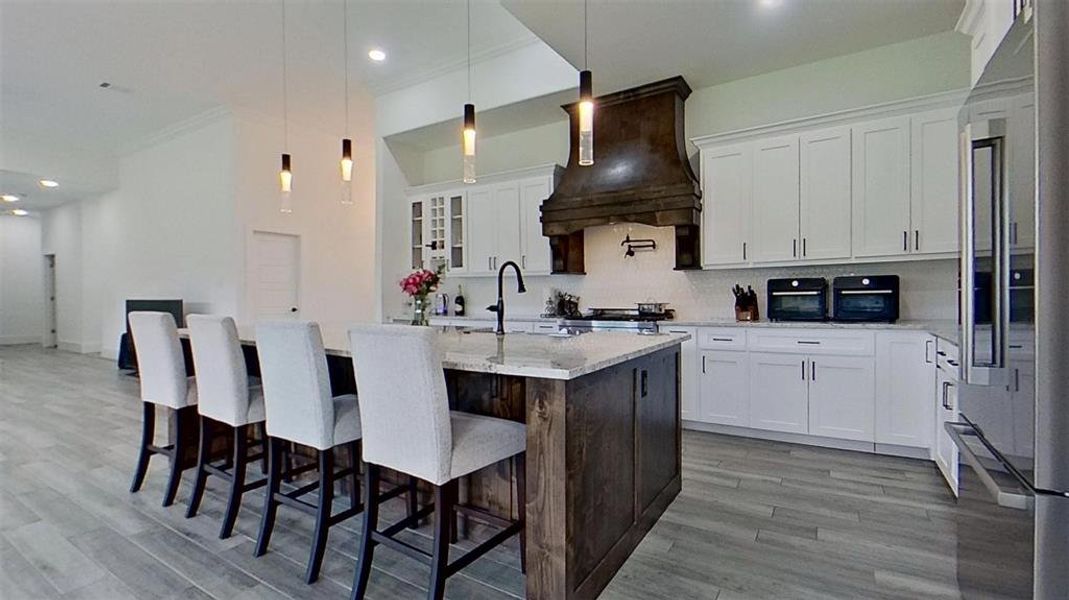 Spacious kitchen featuring a large island with seating, pendant lighting, wood-finish flooring, a decorative range hood, and stainless steel appliances Spacious kitchen featuring a large island with seating, pendant lighting, wood-finish flooring, a decorative range hood, and stainless steel appliances