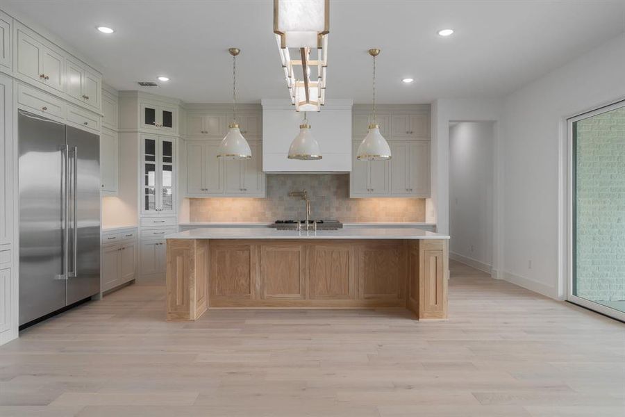 Kitchen featuring built in fridge, tasteful backsplash, pendant lighting, light wood-type flooring, and a kitchen island with sink