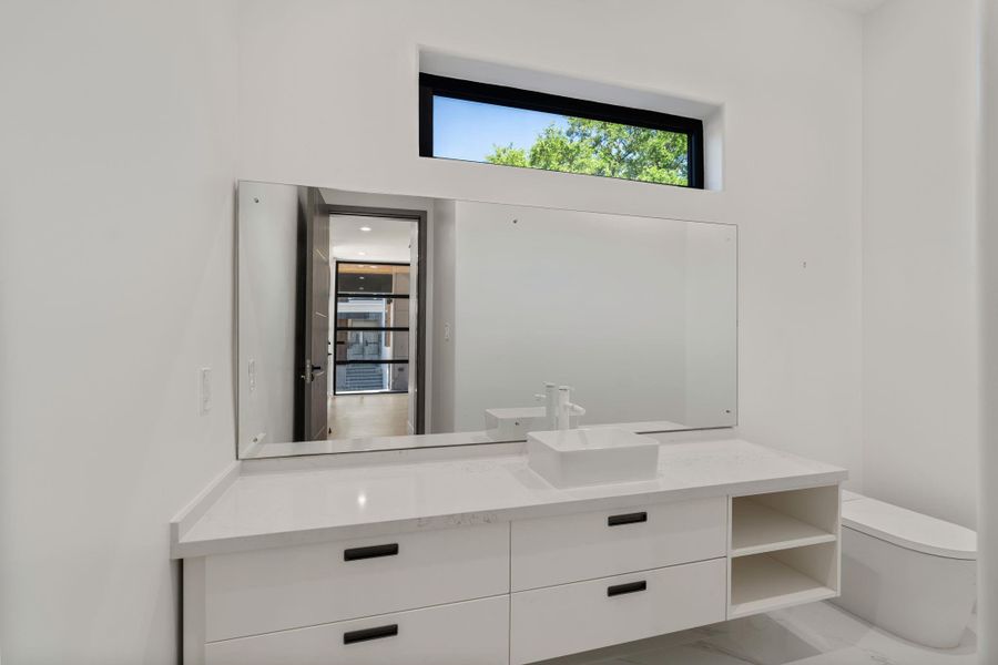 This 1st floor powder bath features a white floating vanity with a vessel sink and black hardware, a full-width frameless mirror, and a narrow horizontal clerestory window that pulls light in from above — a clean, precise finish that holds its own in this meticulously crafted home.