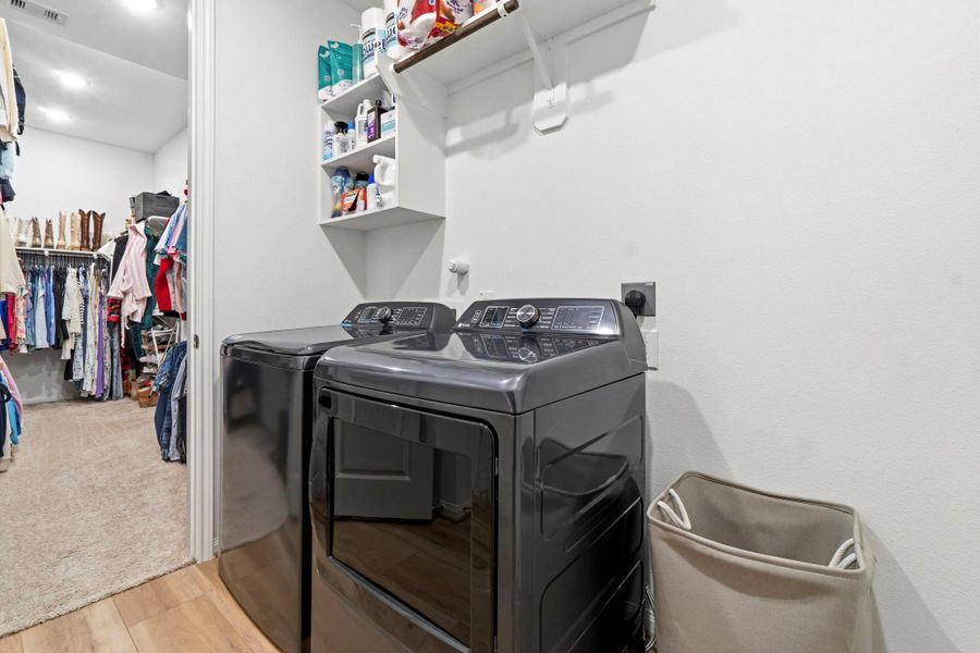 This photo showcases a compact laundry area with modern washer and dryer units, adjacent to a spacious walk-in closet with ample storage and clothing racks leading to the primary bathroom.
