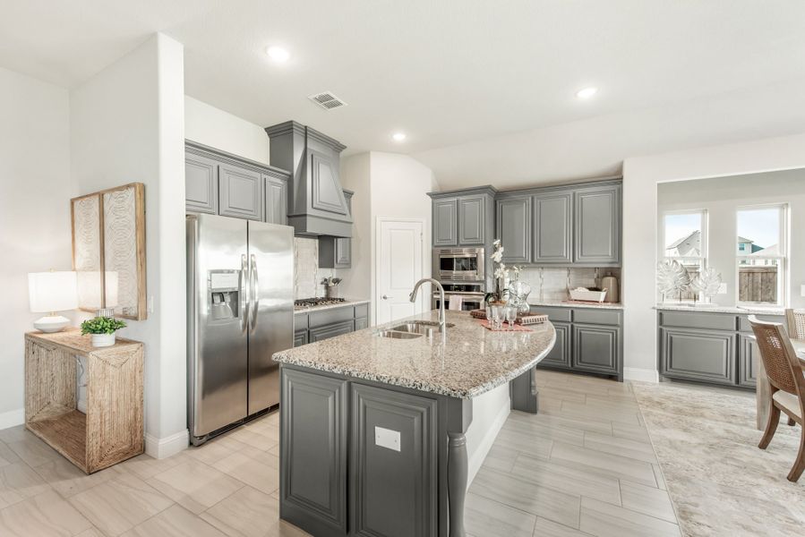 Kitchen with gray cabinets, granite island with sink, and stainless steel appliances on light tile floors