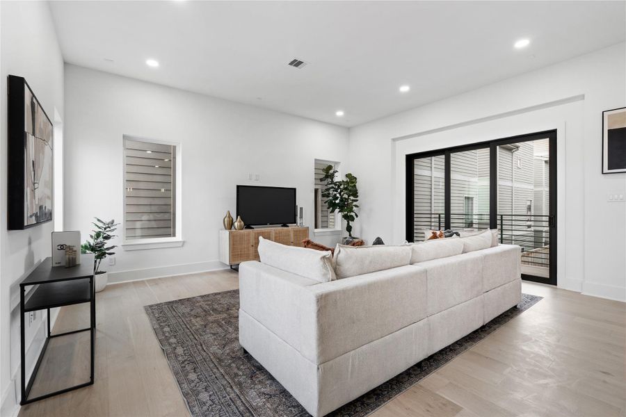 Wrapped in sunlight and anchored by wide-plank flooring, the living area is a serene counterpoint to the home’s bold architectural exterior. Triple-paneled glass sliders frame a covered balcony, blurring the line between indoors and out, while clean walls and natural tones invite personal expression. It's a space designed for slow mornings, lively evenings, and everything in between