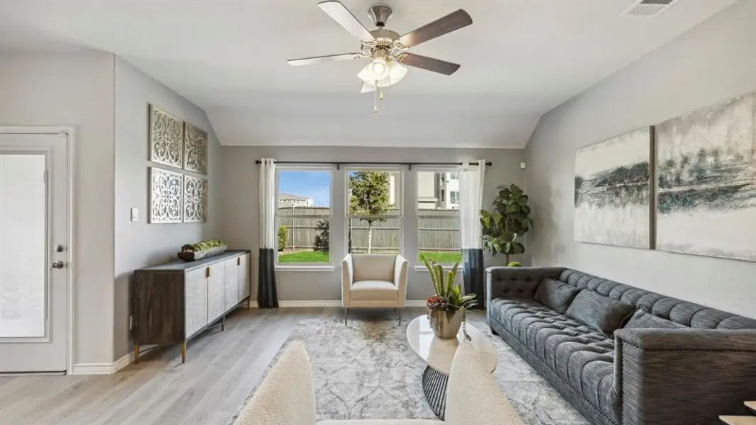 The living area features light-toned flooring, a ceiling fan, and large windows providing views of the green lawn