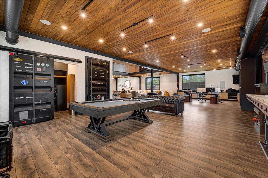 Modern entertainment room features a wooden ceiling and floor, a stylish pool table, and chic seating areas. Large windows offer ample natural light, while the industrial design elements add a contemporary touch. Perfect for relaxation and social gatherings.