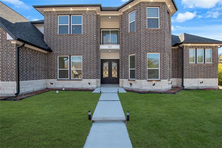 View of front of property with a balcony, a front yard, brick siding, french doors, and stone siding View of front of property with a balcony, a front yard, brick siding, french doors, and stone siding