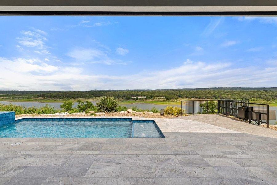 Outdoor living area featuring a rectangular pool with integrated spa, expansive gray-toned paver patio, and black metal railing, all set against a distant water body and natural landscape