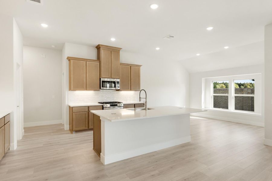 Open kitchen with large white island, tan wood cabinets, stainless appliances, and light hardwood floors Open kitchen with large white island, tan wood cabinets, stainless appliances, and light hardwood floors