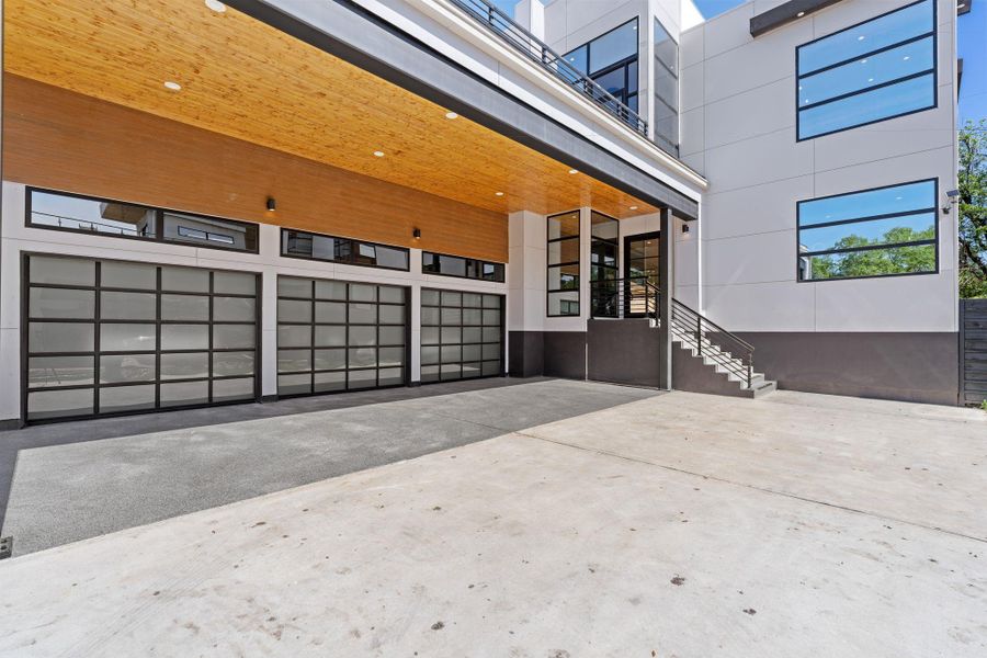 Three striking glass garage bays with black frames line the lower facade beneath the porte-cochere — a covered arrival experience that extends to a staircase leading up to the main entry. The double-wide driveway provides exceptional parking and approach for residents and guests alike.