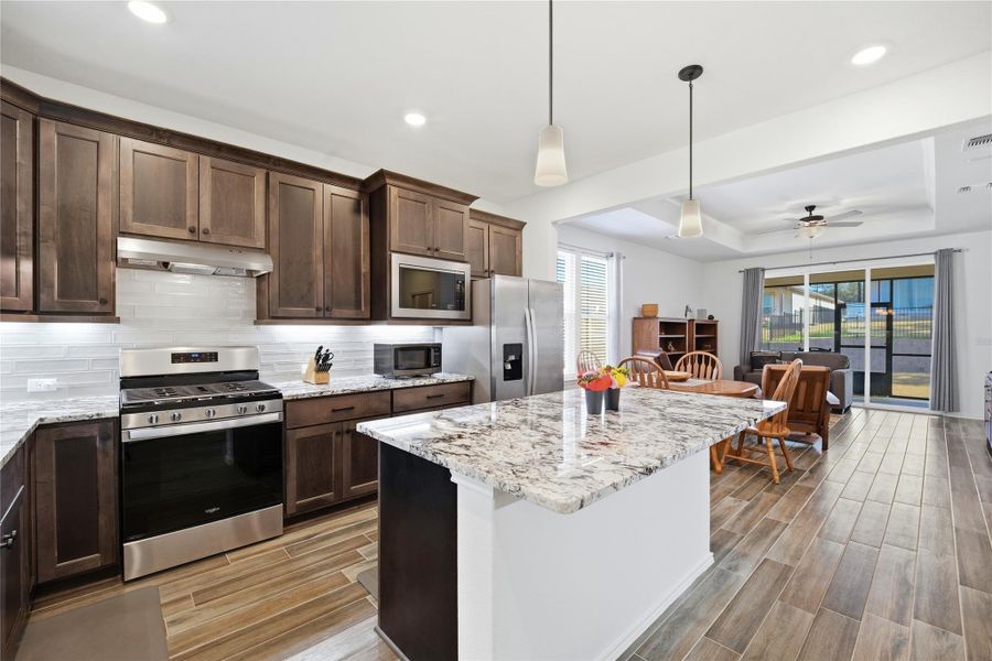 Kitchen with stainless steel appliances, dark wood finish cabinets, pendant lighting, ceiling fan, and light stone countertops