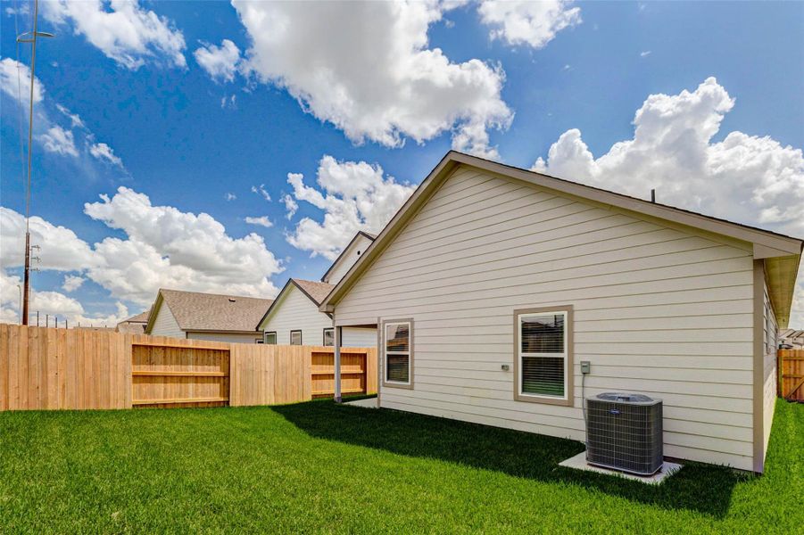 Rear exterior view showcasing the fenced backyard, fresh green lawn, neutral siding, rear windows, and exterior HVAC unit. The private yard offers manageable outdoor space for relaxing, pets, play, or simple weekend enjoyment, with clean new-construction finishes and plenty of sunshine.