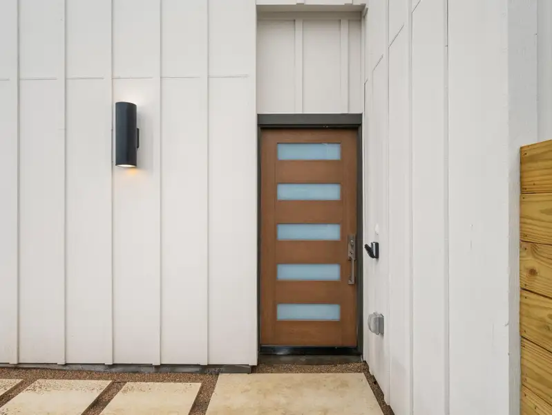 Modern entry featuring a wood-finish door with frosted glass inserts, white board and batten siding, and a contemporary exterior sconce