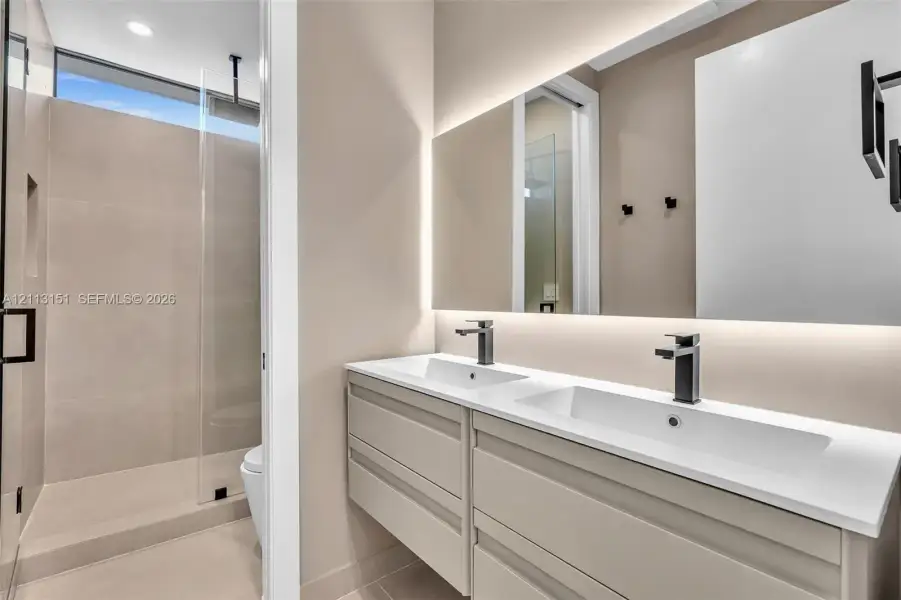 Dual-vanity cabinet with white solid-surface countertop, rectangular undermount sinks, matte black fixtures, and large-format wall mirror with integrated backlighting, featuring a walk-in shower with floor-to-ceiling tile and transom window Dual-vanity cabinet with white solid-surface countertop, rectangular undermount sinks, matte black fixtures, and large-format wall mirror with integrated backlighting, featuring a walk-in shower with floor-to-ceiling tile and transom window
