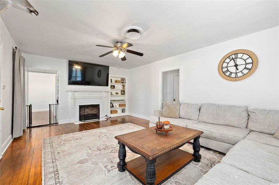 Living room featuring hardwood / wood-style flooring, a fireplace with flush hearth, ceiling fan, and built in features Living room featuring hardwood / wood-style flooring, a fireplace with flush hearth, ceiling fan, and built in features