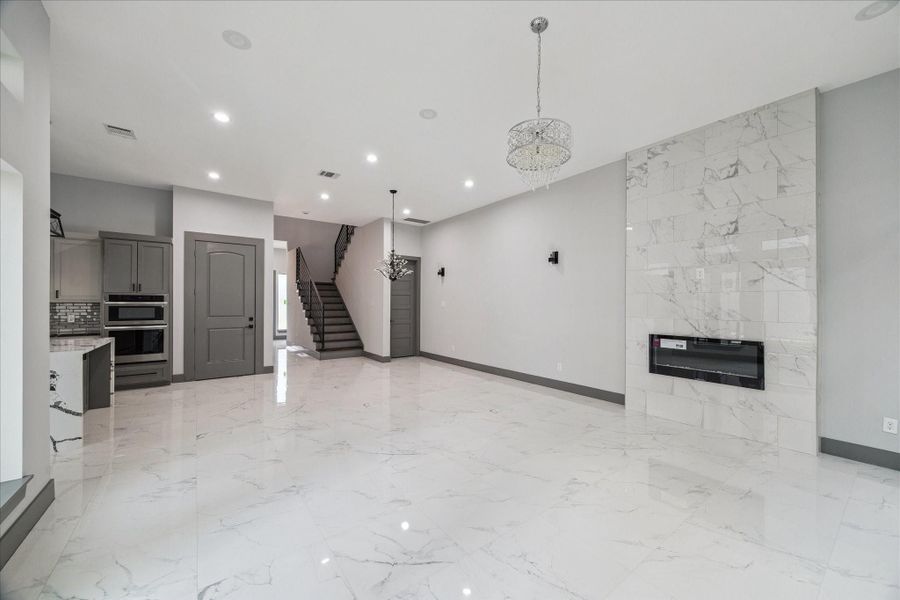 Expansive marble-look tile floors, a sleek modern fireplace, and elegant lighting create a sophisticated setting that flows seamlessly toward the kitchen and staircase. Expansive marble-look tile floors, a sleek modern fireplace, and elegant lighting create a sophisticated setting that flows seamlessly toward the kitchen and staircase.