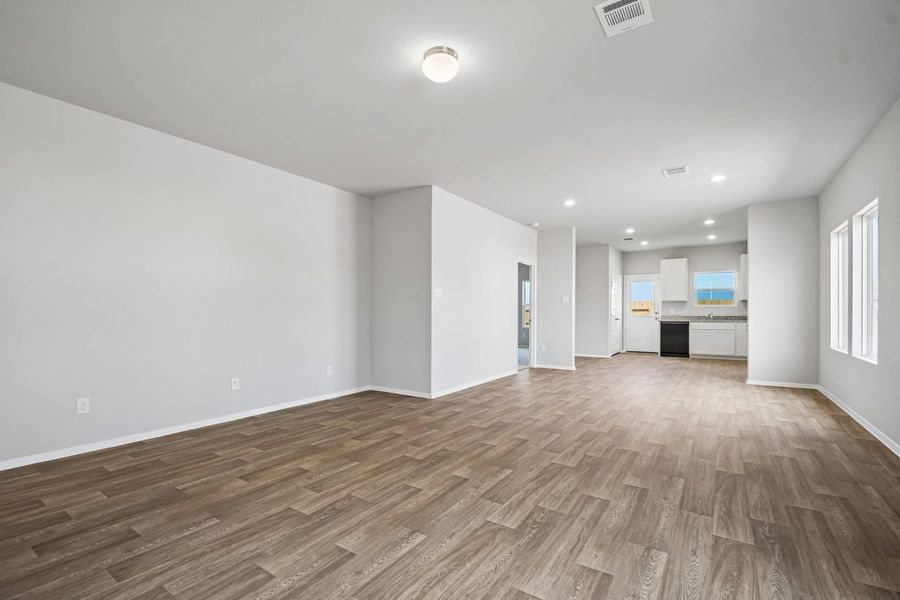 Image of open concept living area in a home with wood-look flooring, white walls, and white cabinets in the kitchen. Windows on the right wall, over kitchen sink, and in the door to back yard.