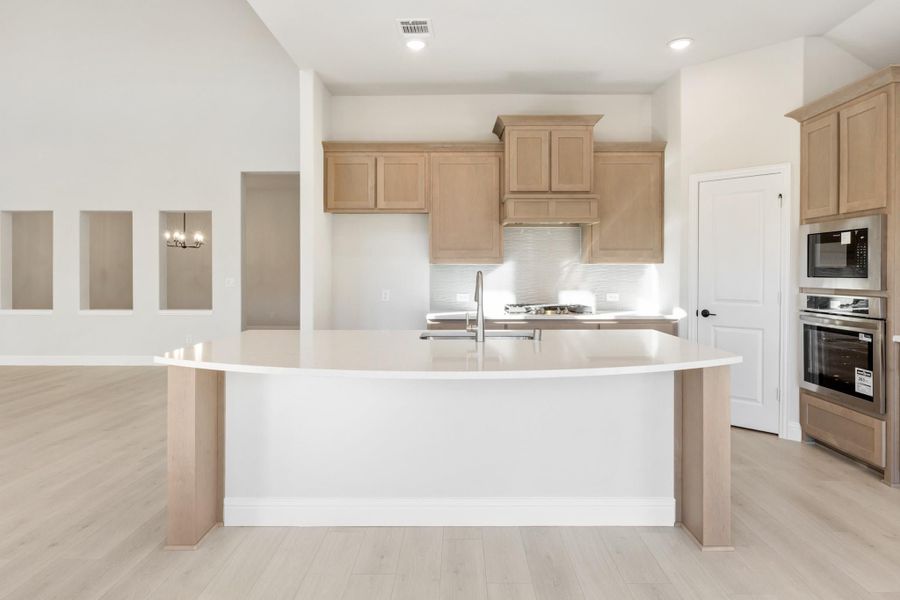 Kitchen with large curved island, light wood cabinets, stainless steel appliances, and light hardwood floors