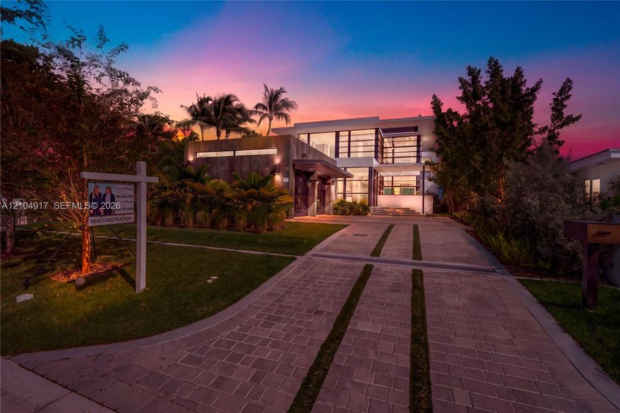 Modern two-story architectural exterior featuring expansive glass windows, stone cladding, a linear paver driveway with integrated turf strips, and mature palm landscaping