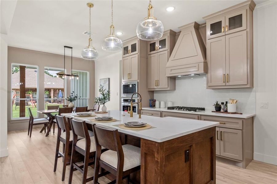 Kitchen featuring custom exhaust hood, appliances with stainless steel finishes, decorative backsplash, light wood finished floors, and an island with sink Kitchen featuring custom exhaust hood, appliances with stainless steel finishes, decorative backsplash, light wood finished floors, and an island with sink