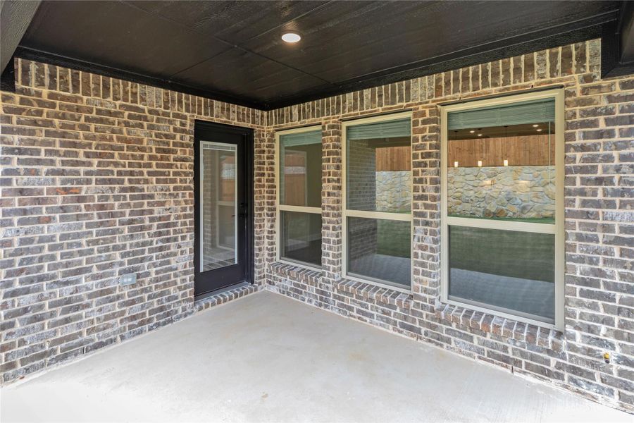 Covered back patio with concrete floor, brick walls, black door, and large windows overlooking interior living area. Covered back patio with concrete floor, brick walls, black door, and large windows overlooking interior living area.