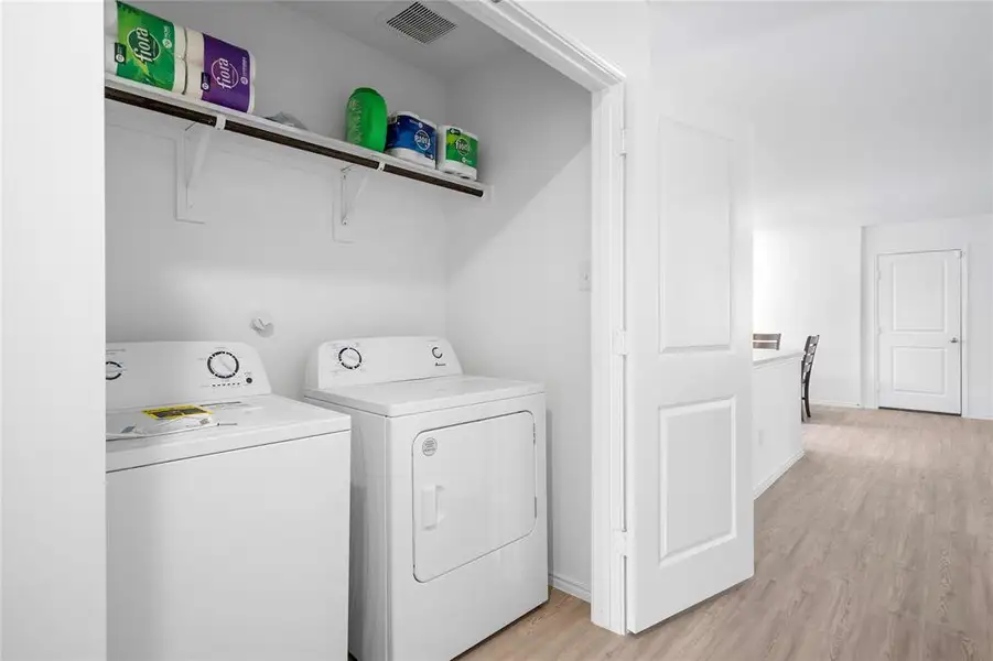 Dedicated laundry closet featuring a built-in shelf, washer and dryer appliances, and light wood-finish flooring extending into the adjacent interior Dedicated laundry closet featuring a built-in shelf, washer and dryer appliances, and light wood-finish flooring extending into the adjacent interior