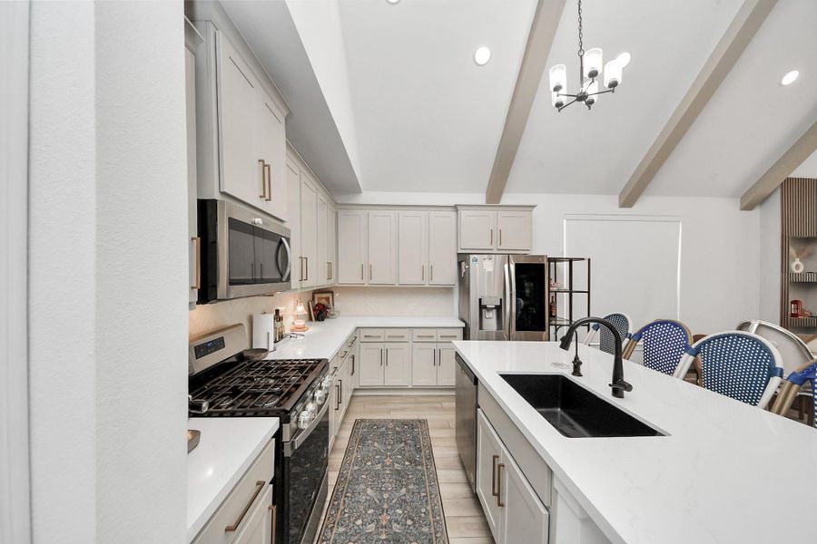This kitchen features modern white cabinetry, stainless steel appliances, and a large central island with a double sink. The space is bright with recessed lighting and a stylish chandelier, complemented by exposed ceiling beams for added character. This kitchen features modern white cabinetry, stainless steel appliances, and a large central island with a double sink. The space is bright with recessed lighting and a stylish chandelier, complemented by exposed ceiling beams for added character.