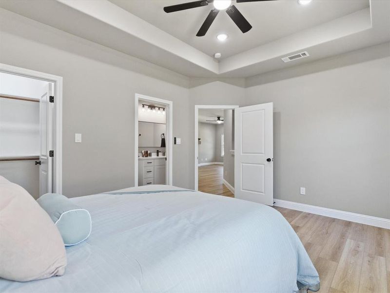 Bedroom featuring light wood finished floors, a raised ceiling, a ceiling fan, ensuite bathroom, and recessed lighting Bedroom featuring light wood finished floors, a raised ceiling, a ceiling fan, ensuite bathroom, and recessed lighting