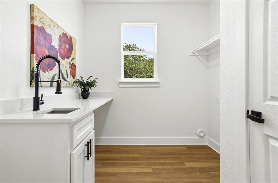 Bright utility room featuring a large window, white cabinetry, a sink with a matte black faucet, white countertops, and wood-finish flooring Bright utility room featuring a large window, white cabinetry, a sink with a matte black faucet, white countertops, and wood-finish flooring