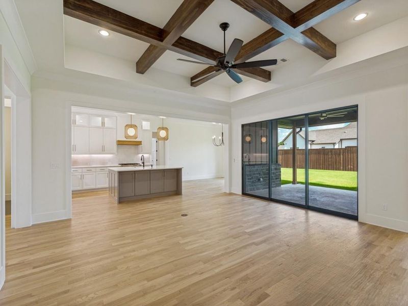 Spacious living area featuring light wood-finish flooring, a coffered ceiling with dark wood beams, and an integrated ceiling fan Spacious living area featuring light wood-finish flooring, a coffered ceiling with dark wood beams, and an integrated ceiling fan